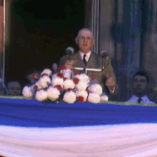 a man in a suit and tie is giving a speech at a podium with flowers in front of him