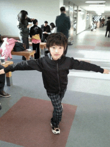 a young boy in a black jacket and plaid pants stands in a hallway with his arms outstretched