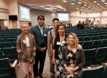 a group of people posing for a picture in a auditorium with a projection screen behind them that says " what are we "