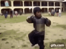 a young boy is running in a field in front of a building while a group of people are standing in the background .