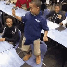 a young boy is standing on a chair in a classroom while a group of children are sitting at tables .