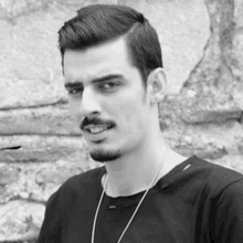 a black and white photo of a young man with a beard wearing a black shirt and a necklace .