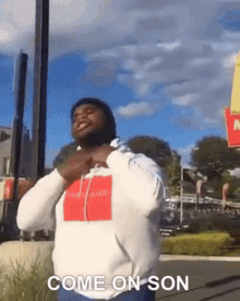 a man in a white hoodie is standing in front of a mcdonald 's and says come on son .