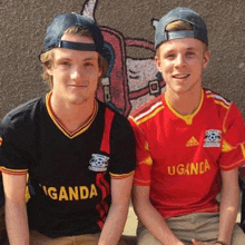 two young men wearing uganda jerseys are sitting next to each other in front of a wall .