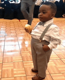 a little boy wearing suspenders and a bow tie holds a hamburger in his hand