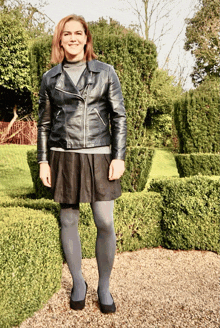 a woman in a black leather jacket and grey tights stands in front of a hedge