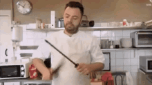 a man in a white shirt is holding a knife in a kitchen with a clock on the wall above him that says nye