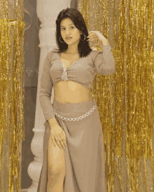 a woman wearing a crop top and a high slit skirt stands in front of a gold curtain