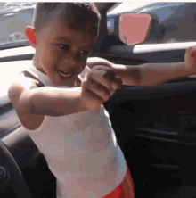 a young boy in a white tank top and red shorts is standing in a car with his arms outstretched