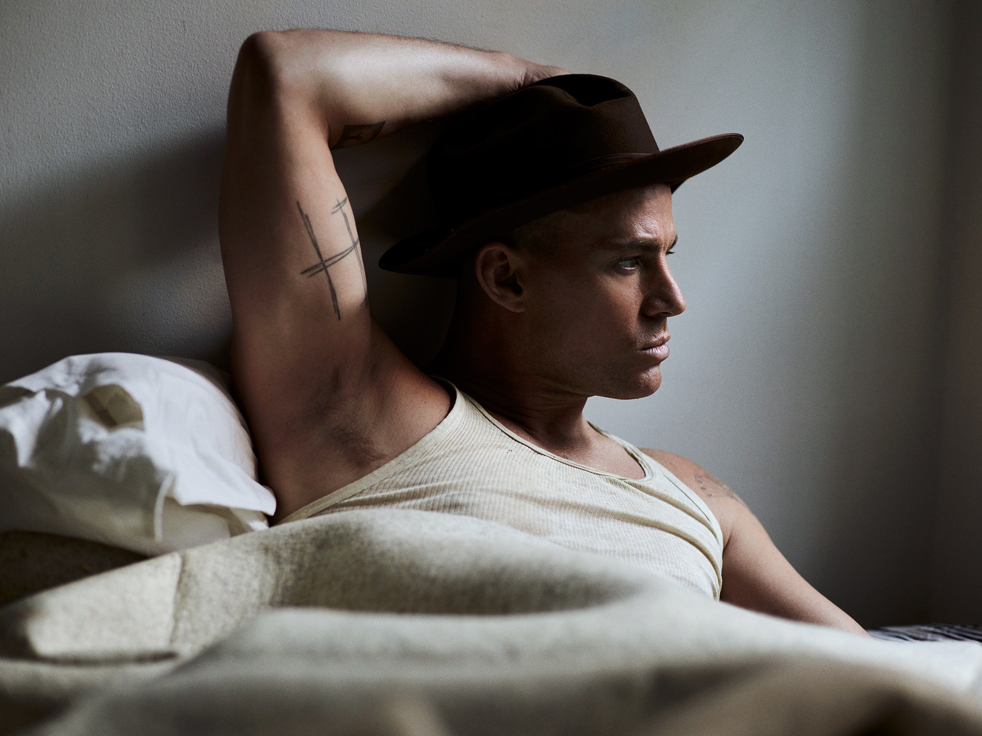 Channing Tatum sits in a bed looking offcamera with his arm above his head. He's wearing a dark cowboy hat and a cream...