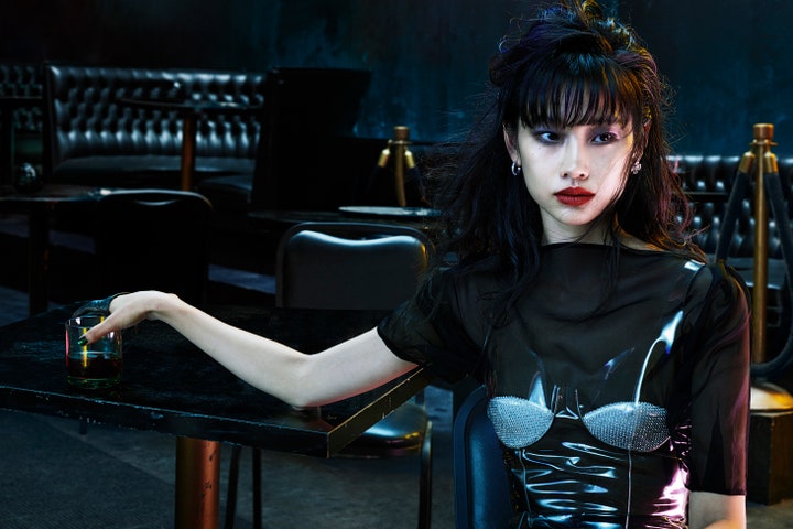 Hoyeon seated at a bar table wearing a black dress by Christopher Kane.