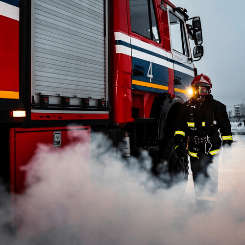 A firefighter in the Netherlands wearing full firefighting gear and standing next to a large, red firetruck.