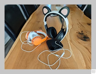 While headphones with cat ears on top hanging from a silver stand with a pair of orange headphones on the table beside it