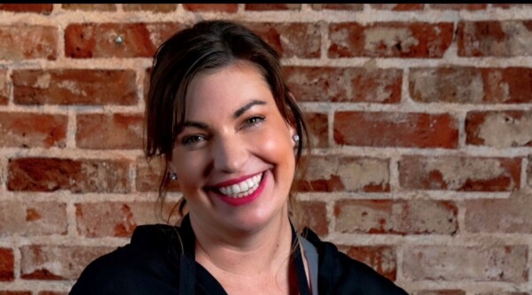 A close-up shot of a woman with brown hair smiling in front of a brick wall.