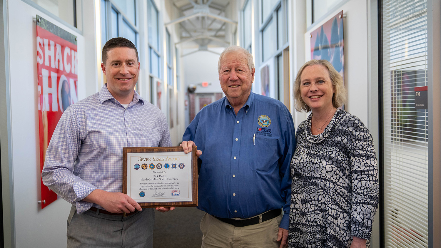 Nick Drake holds a certificate in a hallway, next to two people