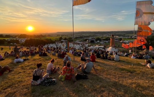 Sunset at the Glastonbury Festival, showing people sitting on a hill looking over the festival