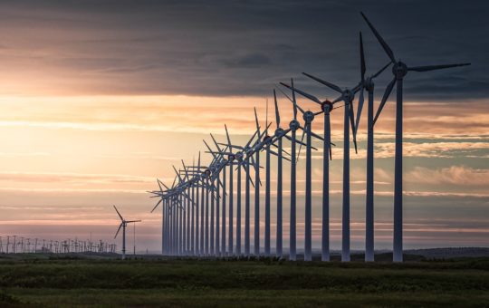 A long line of wind turbines seen against a cloudy sky at dawn or dusk