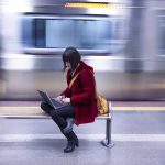 Brand image- Woman in a red coat sitting on a bench working on a laptop in a subway station.