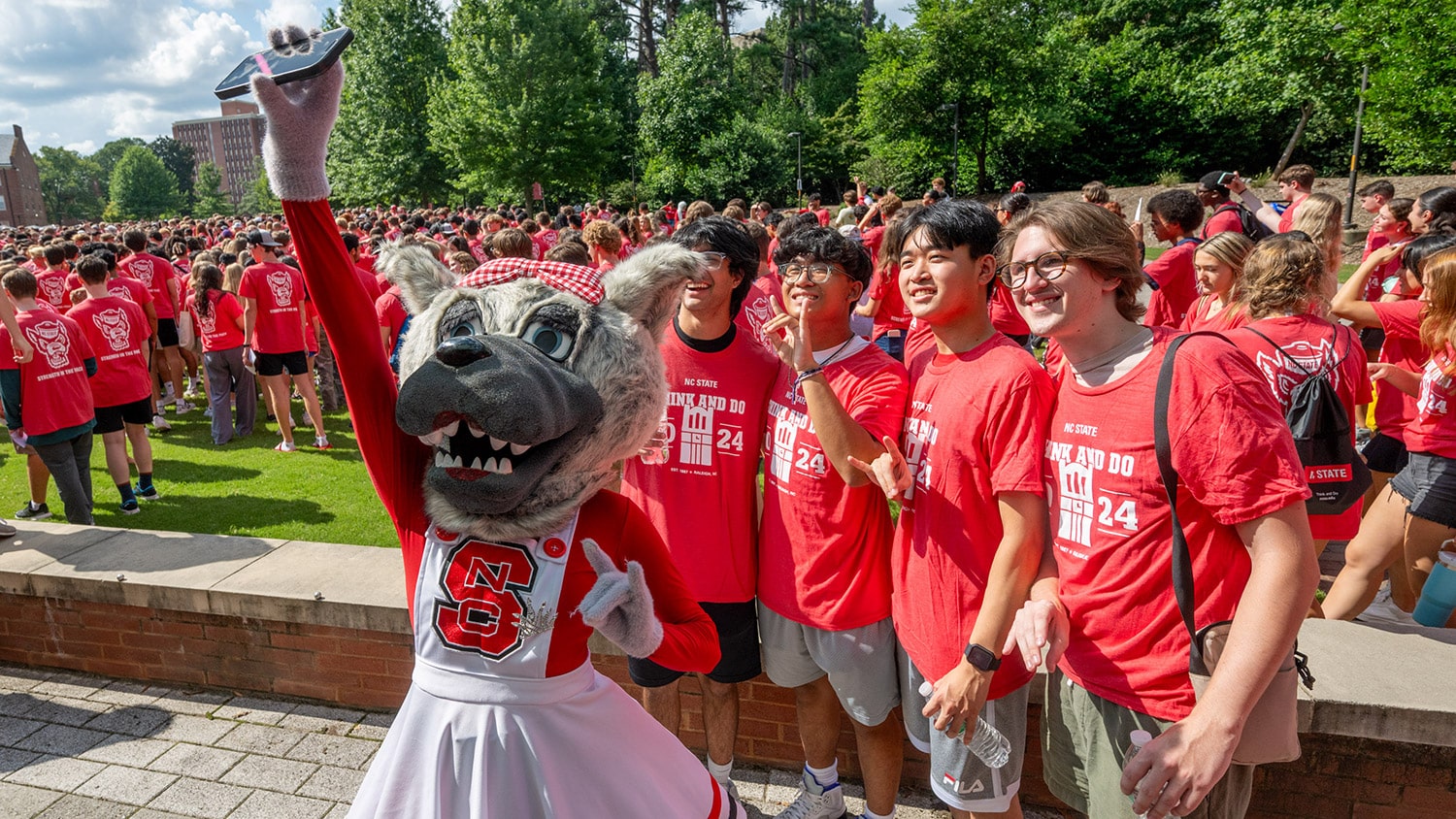 Four students in matching NC State T-shirts pose for a selfie with Ms. Wuf outdoors before taking their class photo during convocation.