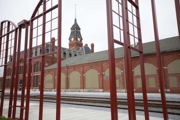 A red metal fence opens to a historic red brick building with a clock tower