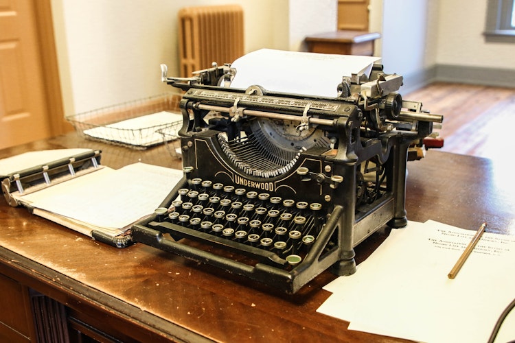 Vintage typewriter and sheets of paper sit on a large wooden desk.