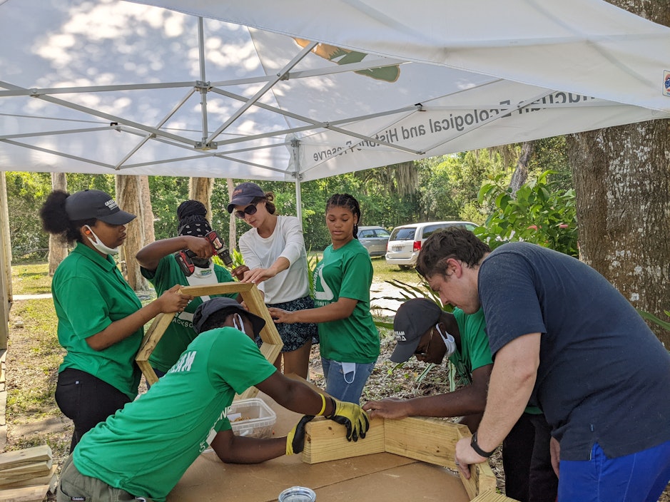 A group of people, working under a pop-up tent, put together a wooden structure