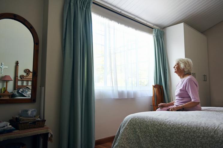 A depressed elderly widow sitting on her bed looking out the window.