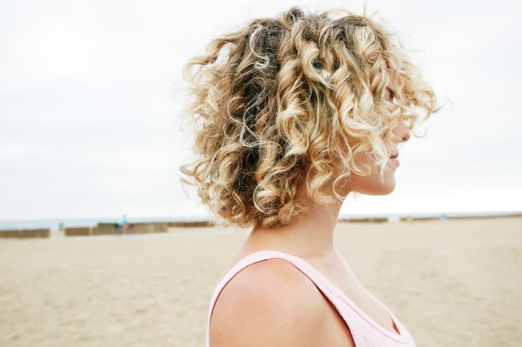 Profile portrait of young woman with blond curly hair standing on sandy beach.