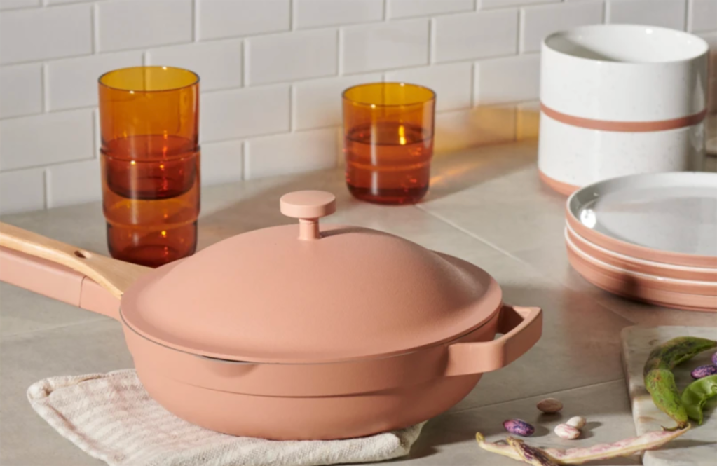 The 'Always Pan' in pink sitting on a counter with Our Place plates and cups