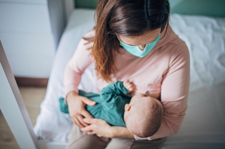 Two people, mother with protective mask breastfeeding her baby son at home, they are at home do to pandemic outbreak quarantine.