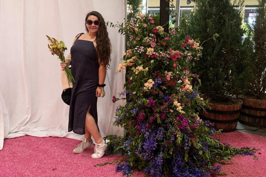Sophie Cannon stands next to a flower arrangement, holding a bouquet and wearing a long dark blue dress with a slit and white sneakers