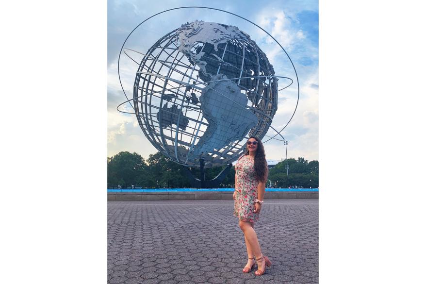 Sophie Cannon stands in a pink floral dress and heels in front of a giant globe at the US Open