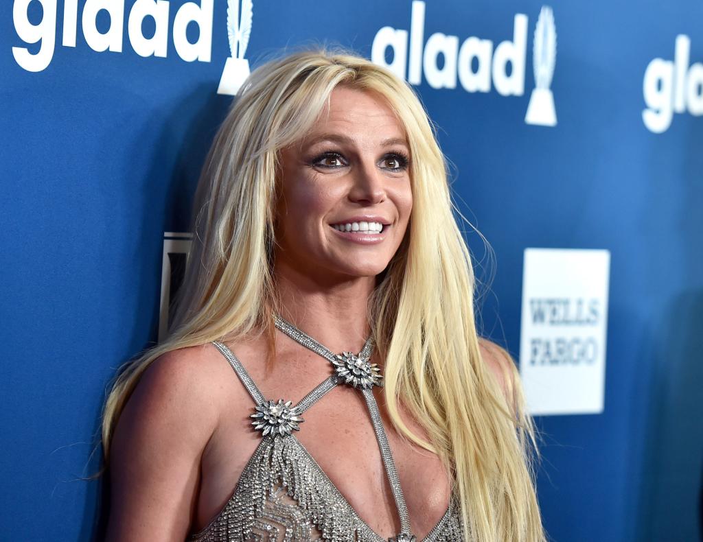 Britney Speras smiles for the cameras wearing a silver dress while standing against a blue backdrop in 2018.