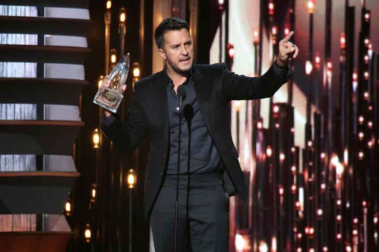 Luke Bryan stands on a stage holding an award and pointing.