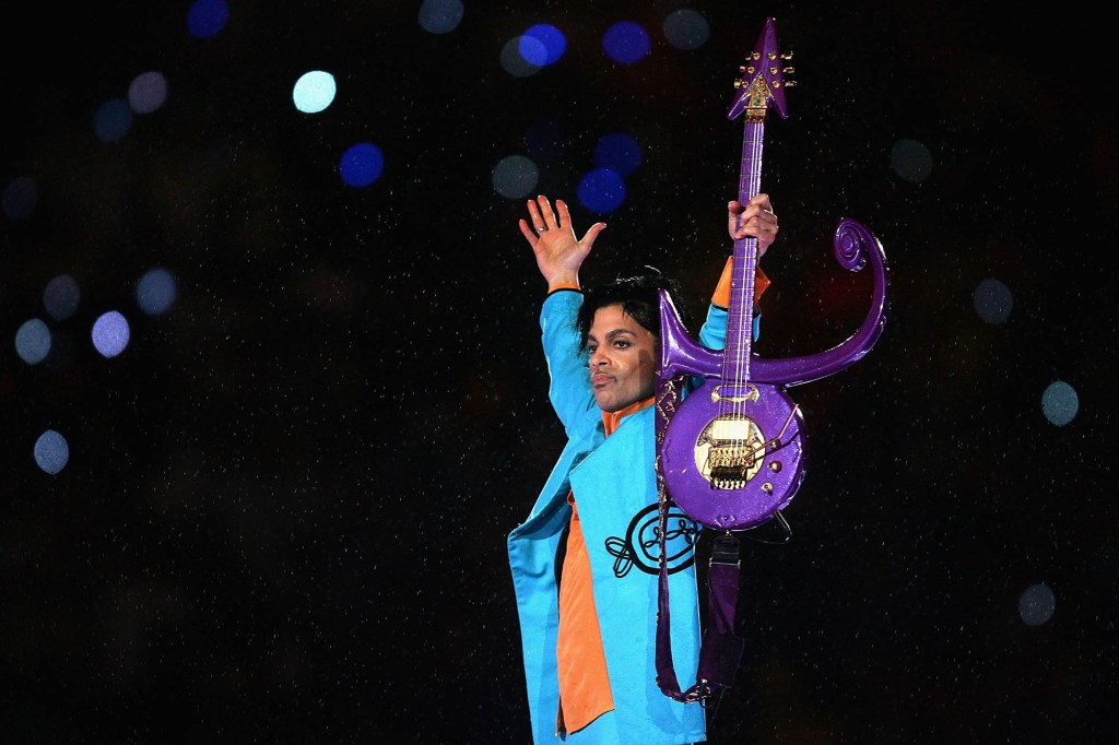 Prince, dressed in blue, holds up his hand and a purple guitar on stage