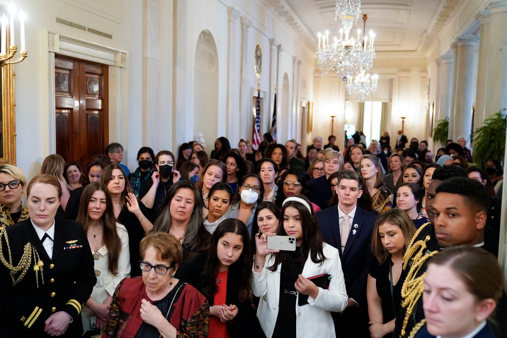 People stand in the Cross Hall of the White House and watch as President Joe Biden speaks during an event to celebrate Equal Pay Day and Women's History Month in the East Room, Tuesday, March 15, 2022.