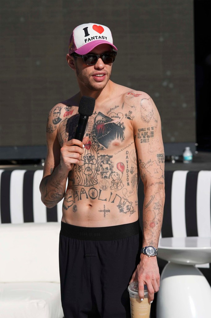 Pete Davidson holds a microphone while shirtless and wearing a trucker hat and black pants.