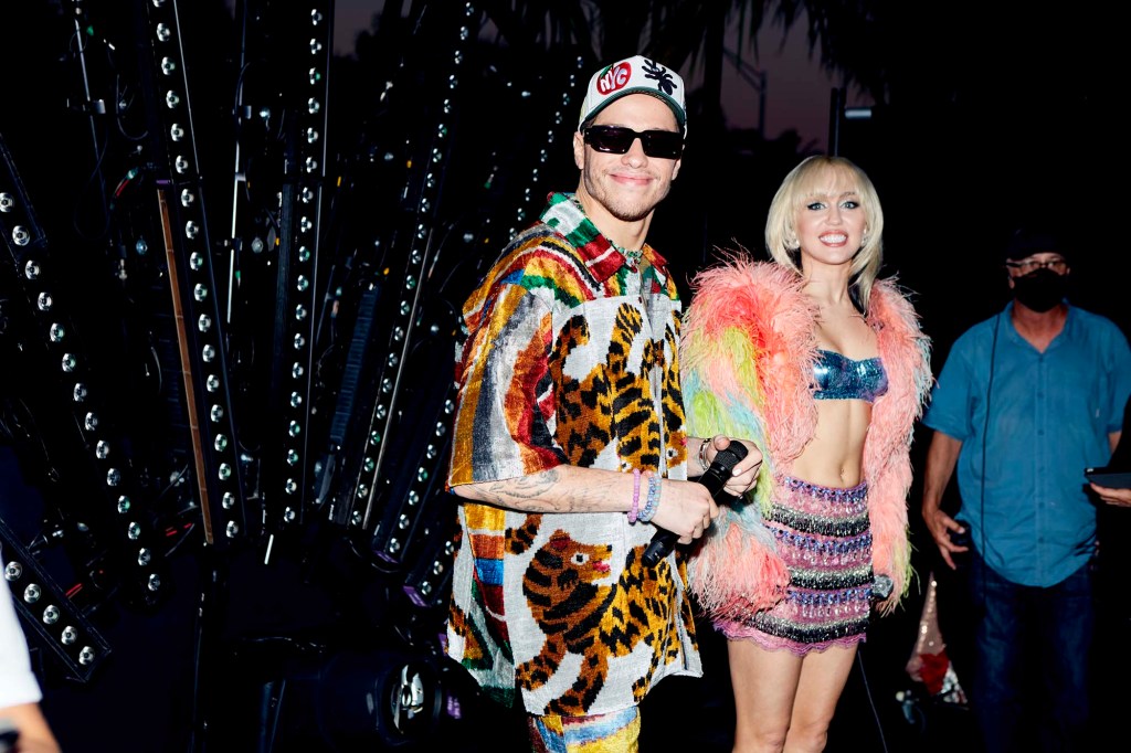 Pete Davidson in a tiger shirt and sunglasses stands next to Miley Cyrus in a fur vest.