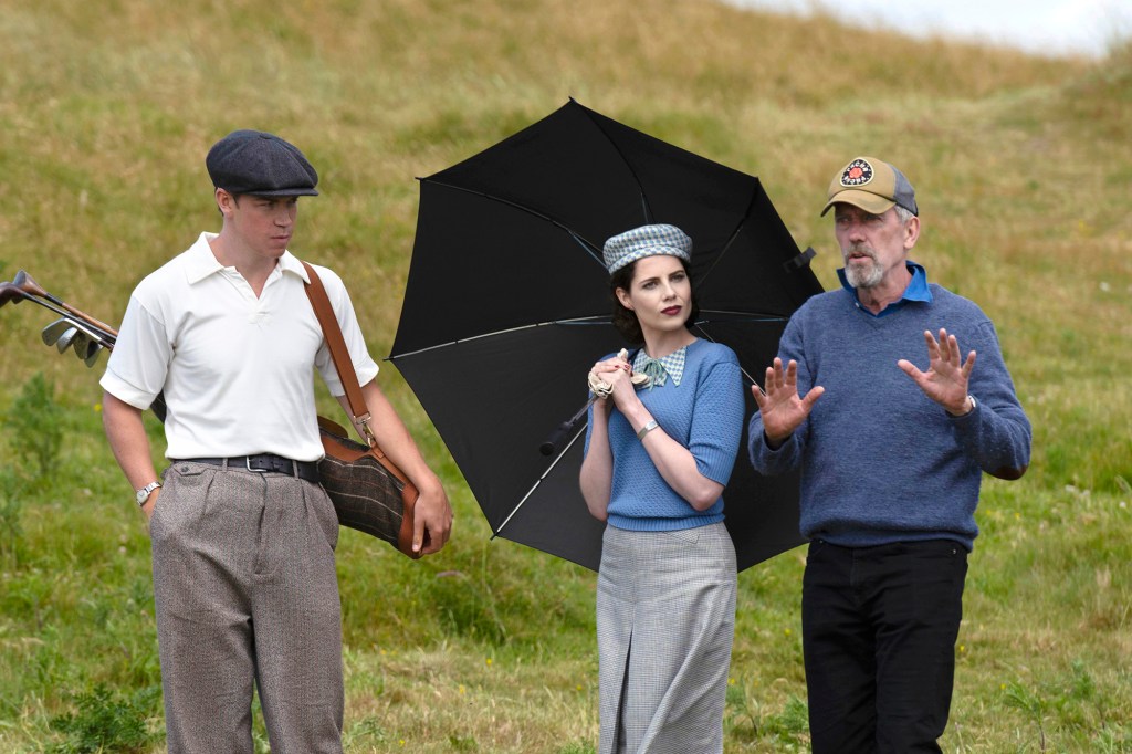 Hugh Laurie directing Will Poulter and Lucy Boynton in a scene from the series. Will is dressed as a caddy and Lucy is holding a black umbrella, which is open. Hugh is wearing a baseball cap, a blue sweater and black pants and is gesturing with his hands.