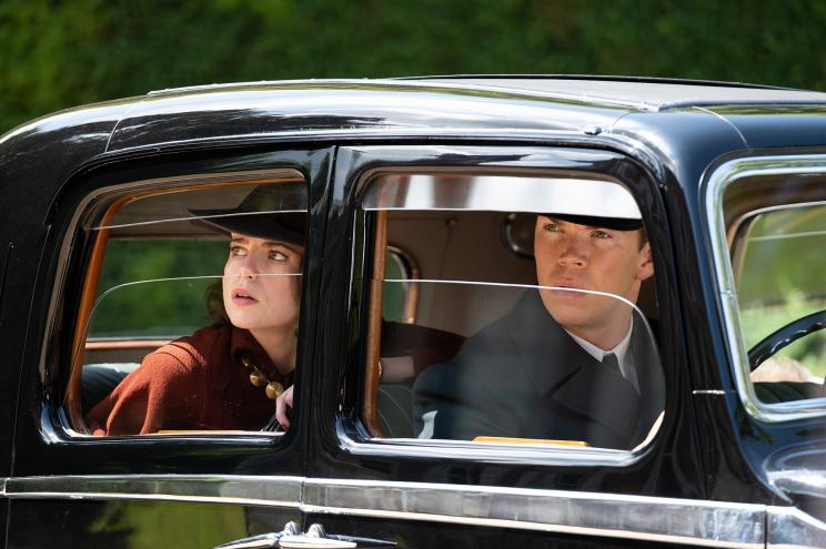 Frankie (Lucy Boynton) and Bobby (Will Poulter) are in disguise and on the case in "Why Didn't They Ask Evans?" Lucy is wearing a black hat and Will is wearing a chauffeur's uniform. They're sitting in a car and looking out the window (Lucy is in the back seat).