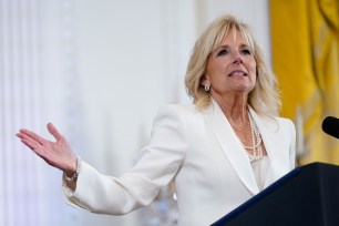 First Lady Jill Biden wearing a white blazer and pearls speaks at a podium.