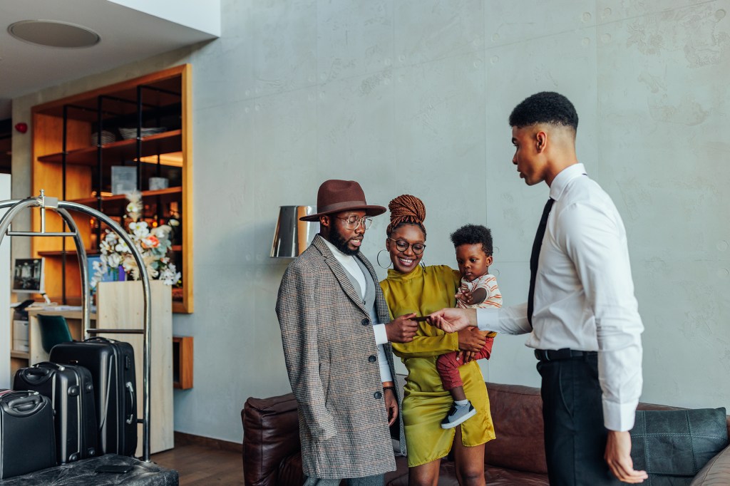 A wealthy African American couple is in the hotel lobby. A young concierge approaches them and is handing the over hotel room key card.