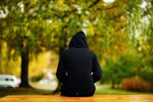 a girl in black hooded shirt with hoodie on her head sitting on a bench not looking at the camera