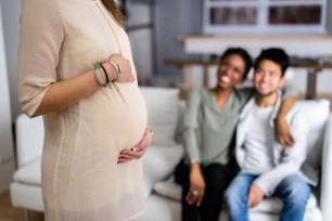 Pregnant Woman Touching Her Belly's Woman Standing In Front Of Smiling Young Couple Sitting