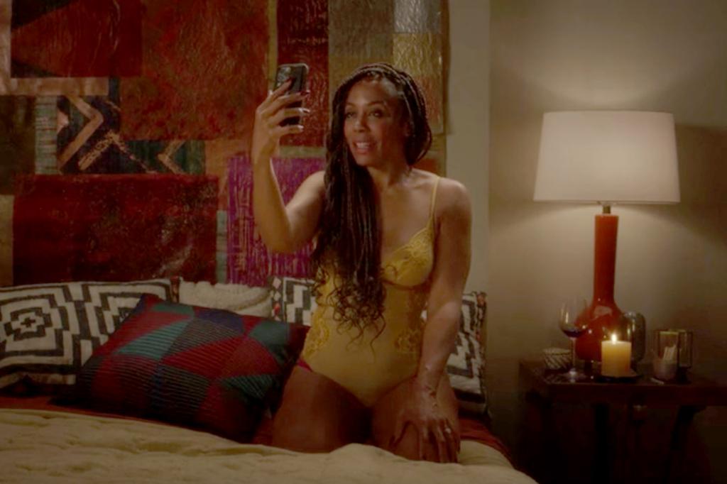 Karen Pittman holds a phone up while on her bed in lingerie. 