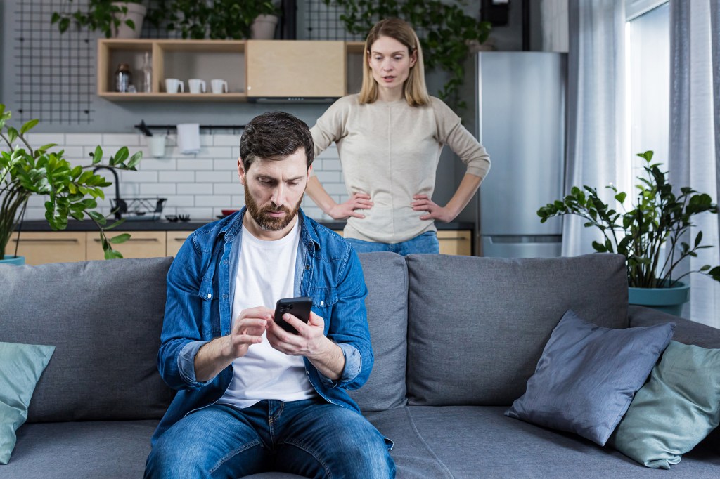 A stock image shot of a husband on his phone while his angry wife stares at him from a distance. 