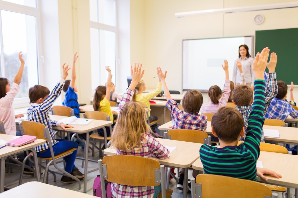 Group of school kids with teacher sitting in classroom and raising hands.