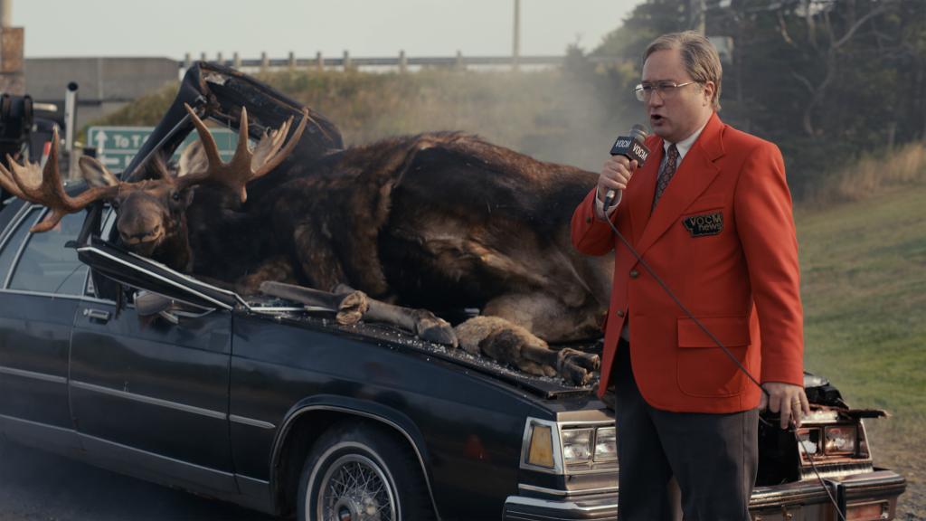 Mark's dad, Mike (Mark Critch) delivers a radio report on a car/moose accident in front of the Critch house, which is next-door to the local radio station. He's wearing a red blazer and is holding a microphone. He's standing in front of a smashed-up car with two dead moose lying where the windshield used to be.