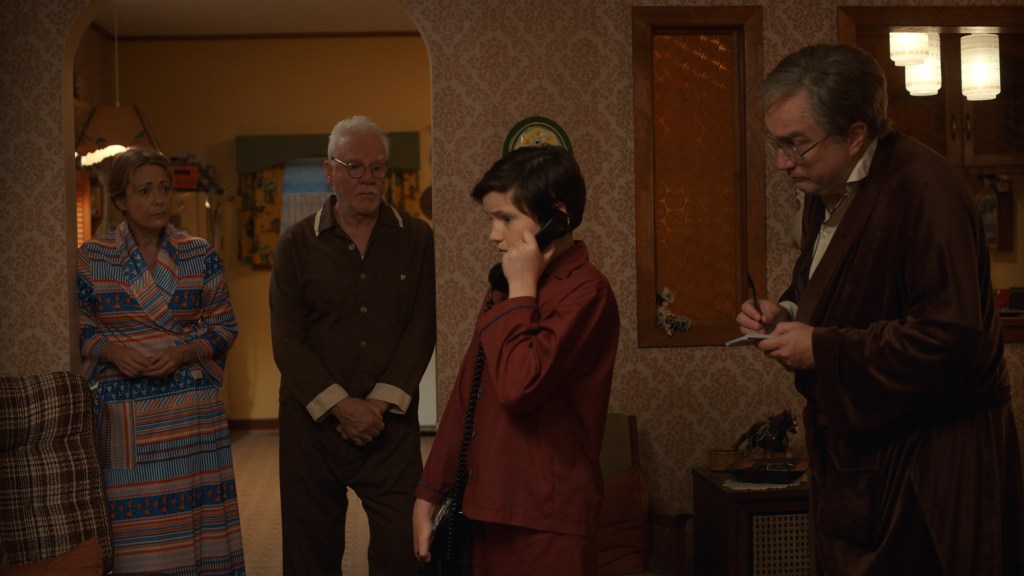 Claire Rankin, Malcolm McDowell, Benjamin Evan Ainsworth and Mark Critch in a scene from "Son of a Critch" on The CW. They're all wearing robes in a scene that appears to be from later in the night in the Critch household. Mark Critch is on the phone.