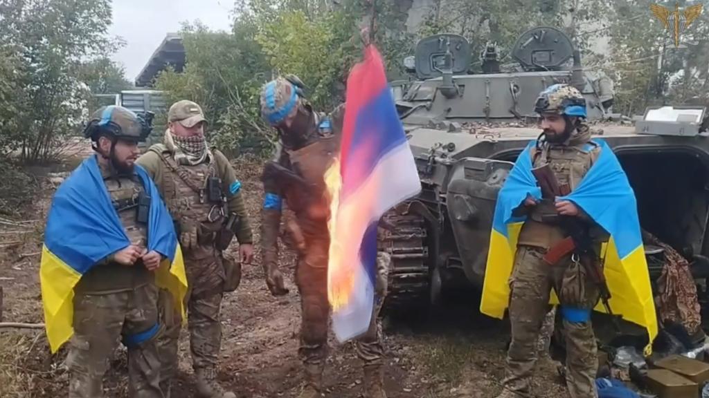 Ukranian soldiers wearing uniforms and wrapped in blue and yellow flags burn a red, white and blue Russian flag while standing on a battlefield nedt to an armored vehicle.